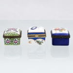 Três(03) caixas porta-joias em porcelana, coleção Porcelain Art. Caixas de pequenas dimensões, destinadas à guarda de anéis, brincos, broches e demais objetos de adorno pessoal, com corpos quadrangulares, tampas abauladas articuladas por dobradiças e guarnições metálicas com fechos frontais. Decoração policromada composta por motivos florais e geométricos, em diferentes combinações cromáticas, com detalhes em douradura. Uma das peças apresenta no reverso marca estampada "Porcelain Art - PA". Medidas: Alt.: 3,5 x Comp.: 4,5 x Larg.: 4cm