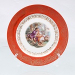 Prato decorativo em porcelana de pasta dura, manufaturado na região da Baviera, Alemanha, durante a segunda metade do século XX. Apresenta formato circular plano com aba larga recoberta por esmalte de tonalidade coral, decorado por gravação contínua em ouro fosco representando crisântemos e ramagens estilizadas. O espelho central exibe vinheta pictórica policromada por processo de decalcomania de alta definição, retratando cena galante clássica ao estilo Fragonard, com casal burguês em trajes do século XVIII dispostos em cenário bucólico arborizado acompanhados de um cordeiro, cercadura interna com guirlandas florais e volutas rococó em ouro polido delimita a transição entre o campo central e a aba, reverso exibe base com anel de assento aparente e selo comercial autocolante ovalado na cor verde, ostentando globo terrestre encimado por coroa e a inscrição "BAVARIA". Medida: Diam.: 17 cm.