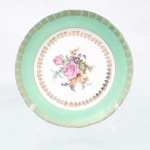 Porcelana Schmidt, Santa Catarina, Brasil (Fundada em 1945). Prato de coleção em porcelana nacional da década de 1950/60. Apresenta formato circular plano com aba larga recoberta por esmalte em tom verde-menta, ornada periféricamente por friso de motivos geométricos gregos repetitivos estampados em ouro, centro exibe arranjo pictórico policromado por processo de decalcomania, composto por buquê de rosas centrais ladeadas por violetas, miosótis e folhagens miúdas, cercadura interna com arabescos e volutas filigranadas em ouro delimita a transição para a aba esmaltada. Marca de manufatura no verso. Medida: Diam.: 17 cm.