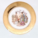 Porcellana Mauá, Mauá - SP(Fundada em 1937 por Fritz Erwin Schmidt / ativa até 1968). Prato de aparato em porcelana nacional, circa 19401950. Apresenta formato circular plano com aba larga integralmente revestida por espessura de ouro, sobre a qual se desenvolve gravação ácida acetinada contendo medalhões rococó, frisos rendados em grega, treliças geométricas e ramagens florais estilizadas, centro exibe composição pictórica policromada sob o tema chinoiserie, retratando duas figuras femininas em trajes tradicionais asiáticos e uma criança junto a balaustrada de jardim sob galhos de cerejeira em flor, ladeados por vaso cerâmico com espécimes botânicos;  cercadura interna com minúsculos florões estilizados em pigmento escuro delimita a transição para a aba dourada. O reverso exibe base com anel de assento aparente, numeração de série "838" pintada à mão livre em pigmento dourado, suporte metálico para suspensão aderido e marca da manufatura. Medida: Diam.: 19,5 cm.