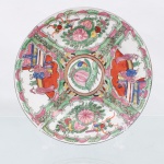 Prato de coleção em porcelana chinesa, concebido no estilo clássico da Família Rosa denominado Medalhão Cantonês. Apresenta organização simétrica com medalhão central circular delimitado por friso dourado e pátina terrosa, contendo a figura de uma ave canora empoleirada sobre folhagem e botão de peônia. Deste núcleo partem quatro reservas radiais ou painéis em formato de leque entrelaçados por borboletas estilizadas e gavinhas em esmalte verde. Dois painéis opostos retratam cenas cortesãs com figuras humanas em terraços arquitetônicos tradicionais, executadas sobre fundo esmalte de tonalidade coral; os dois painéis alternados exibem arranjos naturalistas compostos por peônias, frutos e aves. Reverso apresenta base com anel de assento aparente e marca de atribuição APÓCRIFA quadrangular em sinete vermelho-ferro com quatro caracteres kanji que remetem ao período(Qianlong Nian Zhi / Manufaturado no Reino de Qianlong). Medida: Diam.: 20 cm.