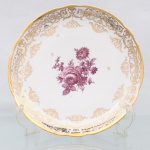 Prato de coleção em porcelana de pasta dura, manufaturado na Europa Ocidental durante o século XIX. Apresenta formato circular com aba ligeiramente recortada em gomos e debrum realçado por filete em ouro, campo central exibe decoração pictórica do tipo "Deutsche Blumen" executada em tonalidade púrpura, representando buquê assimétrico com arranjo de rosas, crisântemos e folhagens detalhadas, aba ostenta rica cercadura ornamental sobreposta em ouro, composta por volutas rococó, ramagens entrelaçadas, cestos suspensos e frisos rendados em guilhoche. O reverso exibe base com anel de assento aparente e medalhão central pintado com botão de rosa em idêntica paleta cromática, ocultando marca de manufatura sob o esmalte. Medida: Diam.: 27 cm.