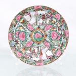 Prato de coleção em porcelana chinesa, seguindo estritamente a gramática ornamental do estilo Medalhão de Cantão da Família Rosa. Superfície interna apresenta organização radial compartimentada a partir de uma reserva central circular circundada por filete dourado, contendo peônia fênix ladeada por ave canora policromada. Da periferia deste núcleo divergem faixas sinuosas que delimitam quatro painéis principais em formato de leque, intermeados por borboletas heráldicas, gavinhas folheadas e botões florais sobrepostos. Duas das reservas opostas abrigam representações minuciosas de cenas palacianas, evidenciando figuras da nobreza em conversação no interior de pavilhões arquitetônicos tradicionais; os dois quadrantes alternados exibem densa composição de peônias abertas, frutos e aves exóticas em ramagens. O reverso apresenta anel de assento plano desprovido de esmalte e sinete APÓCRIFO quadrangular aplicado em pigmento vermelho-ferro sob a inscrição(Qianlong Nian Zhi), alusiva ao reinado da Dinastia Qing. Medida: Diam.: 20,5 cm.