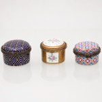 Três(03) caixas porta-joias em porcelana, coleção Porcelain Art. Caixas circulares de pequenas dimensões, destinadas à guarda de anéis, brincos e outros objetos de adorno pessoal, com tampas abauladas articuladas por dobradiças e guarnições metálicas ornamentadas, providas de fechos frontais. Decoração policromada e dourada, duas peças com padrões geométricos reticulados e uma com reservas florais sobre campos em tom ocre. Duas peças conservam no reverso marca estampada Porcelain Art  PA. Medidas: Alt.: 3 x Diam.: 4cm.