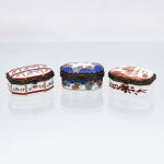 Três(03) caixas porta-joias em porcelana, coleção Porcelain Art. Caixas de formato retangular com cantos chanfrados, destinadas à guarda de anéis, brincos e pequenos objetos de adorno pessoal. Tampas abauladas articuladas por dobradiças, guarnições metálicas ornamentadas e fechos frontais. Decoração policromada: uma com faixas geométricas e motivos florais; outra com campo azul-cobalto reticulado a ouro e cercadura de flores; a terceira com composição monocromática em tom castanho-avermelhado, representando paisagem lacustre com embarcações e vegetação. Uma delas com marca ao fundo "Porcelain Art  PA". Medidas: Alt.: 2,5 x Comp.: 5 x Larg.: 4cm.