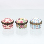 Três(03) caixas porta-joias em porcelana, coleção Porcelain Art. Caixas circulares de pequenas dimensões, destinadas à guarda de anéis, brincos e outros objetos de adorno pessoal, com tampas abauladas articuladas por dobradiças e guarnições metálicas ornamentadas, providas de fechos frontais. Decoração policromada e dourada, sendo uma peça com composição geométrica em tons de rosa, outra com motivos florais em policromia e a terceira com faixas verticais em azul-celeste, rosa e dourado, intercaladas por pequenos elementos florais. Apresentam no reverso marca estampada "Porcelain Art - PA". Medidas: Alt.: 3 x Diam.: 4,5cm.