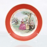 Porcelana D.Pedro II, Rio de Janeiro - RJ(Fundada em 1921 por Tinoco Machado, ativa até a década de 1980). Prato decorativo de parede em porcelana nacional, circa 1950-1960. Esmaltagem vítrea na cor branca, aba larga recoberta por esmalte de tonalidade coral uniforme, delimitada nas extremidades por filetes em ouro  e contornada internamente por cercadura festonada com gavinhas folheadas abstratas. Centro abriga composição pictórica policromada por processo de decalcomania, reproduzindo cena galante de estilo Rococó inspirada nas obras de Jean-Honoré Fragonard. A iconografia retrata figuras da nobreza em trajes do século XVIII dispostas em jardim arborizado, com destaque para duas mulheres tangendo alaúdes ao lado de um homem sentado à mesa de chá, acompanhados por um segundo casal ao fundo à esquerda. A vinheta ostenta as inscrições de atribuição estilística e importação do decalque "FRAGONARD. / M. Langbroek. / Amsterdam. / Holland." no quadrante inferior. Verso exibe base com anel de assento aparente, numeração "212" gravada em baixo-relevo  e marca de manufatura. Medida: Diam.: 23 cm.