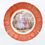 Prato decorativo de parede em porcelana de pasta dura, manufaturado na região da Baviera, Alemanha, durante a segunda metade do século XX. Esmaltagem vítrea na cor branca, aba sutilmente recortada em gomos ondulares e debrum realçado por filete em ouro, recoberta por esmalte de tonalidade coral uniforme, sobreposta por rica ornamentação contínua em ouro fosco composta por volutas acantiformes, buquês e reservas ovais contendo arranjos florais, seguidos por uma cercadura interna festonada em semicírculos filigranados. Centro abriga composição pictórica policromada por processo de decalcomania, retratando cena galante de estilo Rococó com casal da nobreza em trajes do século XVIII executando passos de dança em cenário bucólico arborizado, ladeado à esquerda por músico tocando flauta doce acompanhado por tamboril e, à direita, por dois instrumentistas tangendo violino e flauta transversal próximos a um vaso monumental de jardim. Verso exibe base com anel de assento aparente, etiqueta comercial retangular com inscrição "430 SCP" e selo na cor verde contendo a representação de um globo terrestre encimado por coroa e a inscrição "BAVARIA". Medida: Diam.: 21 cm.