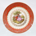Prato decorativo de parede em porcelana de pasta dura, manufaturado na região da Baviera, Alemanha, durante a segunda metade do século XX. Esmaltagem vítrea na cor banca, aba lisa finalizada em debrum dourado, revestida por esmalte de tonalidade coral, sobreposto por gravação contínua em ouro exibindo crisântemos estilizados e folhagens dispostas sobre fundo reticulado em treliça, seguida por uma cercadura interna festonada em semicírculos filigranados. Centro abriga composição pictórica policromada por processo de decalcomania, retratando cena galante de estilo Rococó com casal da nobreza em trajes do século XVIII assentado em gramado sob copa de árvore, com destaque para a figura feminina portando ramalhete de flores junto a uma guirlanda circular disposta ao solo. A vinheta ostenta a assinatura de atribuição estilística "Fragonard" no quadrante inferior direito. Verso exibe base com anel de assento aparente, e selo comercial ovalado na cor verde, contendo a representação de um globo terrestre encimado por coroa, a inscrição "BAVARIA"(marca de sobreposição" e o dístico inferior "FAVORIT". Medida: Diam.: 19,5 cm.