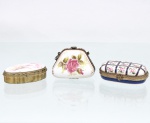 Três(03) caixas porta-joias em porcelana, coleção Porcelain Art. Composto por caixas de pequenas dimensões, em formatos oval, trapezoidal e oblongo, destinadas à guarda de joias e pequenos objetos pessoais. Confeccionadas em porcelana com tampas articuladas, guarnições metálicas ornamentadas e fechos frontais; o exemplar trapezoidal possui corrente metálica lateral, permitindo também seu uso como pequena bolsa de mão. Decoração policromada predominantemente floral, com rosas e folhagens, complementada por filetes e detalhes em douradura; uma das peças apresenta composição reticulada em azul-cobalto com reservas florais. Apresentam marca estampada "Porcelain Art - PA". Medidas: Alt.: 3,5 x Comp.: 5 x Larg.: 3cm.Medidas: Alt.: 2 x Comp.: 5,5 x Larg.: 3cm.