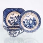 Trio para chá em faiança japonesa, Imperial Ironstone Ware, N.K.T., Japão, meados do século XX. Composto por xícara, pires e prato de sobremesa quadrangular com cantos arredondados. Decoração derivada do Willow Pattern, em azul sob esmalte, com paisagem orientalista constituída por arquitetura, ponte, arvoredo, curso d'água, embarcação e aves em voo; cercaduras geométricas em azul e sépia e filetação dourada. Superfície com craquelê de esmalte. No reverso, marca "Imperial Ironstone Ware - N.K.T. - Made in Japan", vinculada à produção da Nihon Koshitsu Toki, atual NIKKO, manufatura fundada em Kanazawa em 1908. Medidas xícara: Alt.: 5,5 x Comp.: 10 x Larg.: 8cm.Medidas pires: Diam.: 13,5cm.Medidas prato: Comp.: 14 x Larg.: 14cm.