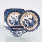 Trio para chá em faiança japonesa, Imperial Ironstone Ware, N.K.T., Japão, meados do século XX. Composto por xícara, pires e prato de sobremesa quadrangular com cantos arredondados. Decoração derivada do Willow Pattern, em azul sob esmalte, com paisagem orientalista constituída por arquitetura, ponte, arvoredo, curso d'água, embarcação e aves em voo; cercaduras geométricas em azul e sépia e filetação dourada. Superfície com craquelê de esmalte. No reverso, marca "Imperial Ironstone Ware - N.K.T. - Made in Japan", vinculada à produção da Nihon Koshitsu Toki, atual NIKKO, manufatura fundada em Kanazawa em 1908. Medidas xícara: Alt.: 5,5 x Comp.: 10 x Larg.: 8cm.Medidas pires: Diam.: 13,5cm.Medidas prato: Comp.: 14 x Larg.: 14cm.