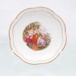 Prato decorativo de parede em porcelana de pasta dura, manufaturado na região da Baviera, Alemanha, durante a segunda metade do século XX. Esmaltagem vítrea na cor branca, aba moldada em gomos ondulares, caracterizada por cercadura periférica com galerias de pequenos orifícios retangulares vazados(reticulated porcelain), finalizada em debrum superior e inferior com filetes em ouro polido. Centro abriga composição pictórica policromada por processo de decalcomania de inspiração neoclássica, retratando três figuras femininas com vestes de estilo helenístico dispostas em cenário campestre arborizado, ladeadas à direita por ramagens de roseiras em flor e, à esquerda, por uma das figuras executando acordes em uma lira de braço. Verso exibe base com anel de assento aparente, numeração "8" pintada sob o esmalte, etiqueta comercial retangular com inscrição "1552 KP" e marca de manufatura constituída por coroa real encimando o dístico 'Bavaria". Medida: Diam.: 15,5 cm.