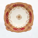 NORITAKE, Nagoya - Japão(Fundada em 1904 pelos irmãos Ichizaemon e Toyo Morimura). Bolw executado em porcelana japonesa,  datável de meados do século XX. Apresenta formato hemisférico, bordas recortadas em ondulações suaves e debrum realçado por filete dourado e cantos arredondados. A ornamentação exibe proeminente faixa perimetral em esmalte vermelho-rubi ou borgonha, sobreposta por densa folhagem e corolas florais estilizadas em douradura aplicada. Esta cercadura encarnada é emoldurada por ampla moldura externa e filetes internos em tom dourado opaco com textura em relevo miúdo mimetizando grânulos ou arabescos entrelaçados. Centro, de fundo esbranquiçado e liso, ostenta medalhão circular simétrico composto por uma roseta de crisântemo circundada por volutas e ramagens fitomórficas neoclássicas em tom ouro. Marca da manufatura com o carimbo oficinal dourado sob o vidrado, acompanhada pela inscrição arqueada inferior "MADE IN JAPAN". Medidas: Alt.: 5 x Diam.: 17cm.
