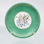 Prato decorativo de parede em porcelana nacional, circa 1960-1970. Esmaltagem vítrea na cor branca, aba larga revestida por esmalte verde-esmeralda acetinado, ornada por debrum periférico contendo friso de espigas estilizadas e motivos fitomorfos heráldicos em ouro, seguidos por uma cercadura interna recortada em arabescos e volutas metalizadas. Centro abriga arranjo pictórico policromado executado à mão livre sobre o fundo branco, representando buquê naturalista com rosa centralizada, miosótis, flores campestres e folhagens detalhadas. A composição ostenta a assinatura "H.B. Curty". Verso exibe base com anel de assento aparente e marca de manufatura. Medida: Diam.: 18,5cm.