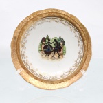 Bowl decorativo em porcelana nacional, circa 1960-1970. Formato hemisférico raso, esmaltagem vítrea na cor branca, aba moldada em gomos ondulares suaves, integralmente revestida por espessa camada de ouro, sobre a qual se desenvolve gravação contínua acetinada composta por crisântemos abertos e ramagens estilizadas, cercadura interna filigranada em ouro com motivos florais e volutas delimita a transição entre a aba e o centro. Este abriga composição pictórica policromada por processo de decalcomania, representando cena galante de estilo Rococó com silhuetas de um casal em trajes da nobreza do século XVIII executando passos de dança sobre piso axadrezado, ladeados por muretas de jardim sustentando vasos monumentais com roseiras e arbustos topiados ao fundo. Verso exibe base com anel de assento e marca de sobreposição em dourado. Medidas: Alt.: 3,5 x Diam.: 14,5cm.