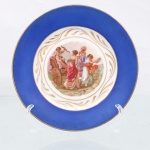 Porcellana Mauá, Mauá - SP(Fundada em 1937 por Fritz Erwin Schmidt / ativa até 1968). Prato decorativo de parede em porcelana nacional, circa 19401950. Esmaltagem vítrea na cor branca, aba larga revestida por esmalte azul-cobalto fosco, delimitada na extremidade periférica por filete em ouro; cercadura interna sobre o fundo branco exibe guirlanda contínua de folhas e gavinhas estilizadas em douradura, que delimita a transição para centro. Este abriga composição pictórica policromada por processo de decalcomania de inspiração neoclássica, baseada nas obras da pintora suíça Angelica Kauffman. A iconografia retrata Cupido(Eros) alado empunhando arco e assentado sobre uma biga de madeira, ladeado por três figuras femininas em trajes de época espalhando flores em cenário campestre. A vinheta ostenta a assinatura de atribuição estilística "Angelica Kauffman" no quadrante inferior esquerdo. Verso exibe base com anel de assento aparente, carimbo de decoração ovalado em pigmento vermelho-ferro contendo a inscrição "ATELIER A. BRANCA" e marca de manufatura. Medida: Diam.: 17cm.