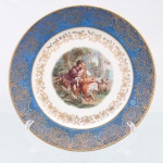 Prato decorativo em porcelana de pasta dura, manufaturado na região da Baviera, Alemanha, durante a segunda metade do século XX. Esmaltagem vítrea na cor branca, aba recoberta por esmalte de tonalidade azul-celeste uniforme, sobreposta por rica ornamentação contínua em ouro composta por crisântemos estilizados e detalhamento rendado em padrão guilhoché, seguida por uma cercadura interna festonada contendo ramagens e corolas florais em douradura. Centro abriga composição pictórica policromada por processo de decalcomania, retratando cena galante de estilo Rococó com casal de pastores em cenário bucólico arborizado com cercado de madeira, ladeado à direita por uma ovelha e, à esquerda, por um cesto com rosas. Verso exibe base com anel de assento aparente, selo na cor verde contendo a representação de um globo terrestre encimado por coroa com a inscrição "BAVARIA" e o número "17". Medida: Diam.: 17 cm.