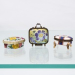 Três(03) caixas porta-joias em porcelana, coleção Porcelain Art. Peças de pequenas dimensões, destinadas à guarda de joias e pequenos objetos pessoais, com tampas articuladas, montagens metálicas e fechos frontais. Compreende caixa oval com decoração policroma de rosas; caixa quadrangular em formato de pequena valise, com alça metálica e composição floral em tons de violeta sobre fundo amarelo; e caixa oval elevada sobre quatro pés, decorada em azul-cobalto, vermelho e dourado. Apresentam na base a marca "Porcelain Art - PA". Medidas: Alt.: 3 x Comp.: 5 x Larg.: 4,5cm.Medidas: Alt.: 2,5 x Comp.: 6 x Larg.: 5cm.Medidas: Alt.: 2 x Comp.: 5,5 x Larg.: 5,5cm.