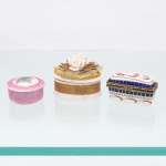 Três(03) caixas porta-joias em porcelana, coleção Porcelain Art e outras manufaturas. Peças de pequenas dimensões, destinadas à guarda de joias e pequenos objetos pessoais. Compreende caixa circular em tonalidade rosa, com tampa removível decorada por composição de violetas e ornatos dourados, trazendo inscrições e numeração no reverso; caixa oval em porcelana branca, circundada por faixa dourada com arabescos, tampa ornamentada com rosa modelada em relevo, folhas e guarnição têxtil com contas douradas; e caixa retangular Porcelain Art, com tampa articulada, montagens metálicas douradas, fecho floral e decoração policroma composta por reservas florais, frisos azul-cobalto, ramos verdes e filetes dourados. Marca "Porcelain Art  PA" no reverso da última peça. Medidas: Alt.: 4,5 x Comp.: 6 x Larg.: 4cm.Medidas: Alt.: 3,5 x Comp.: 5,5 x Larg.: 3,5cm.Medidas: Alt.: 2,5 x Diam.: 4,5cm.