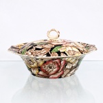 Myott Son & Co., Staffordshire, Inglaterra(Fundada em 1898). Terrina coberta em faiança inglesa, datável do período compreendido entre as décadas de 1930 e 1940. Pertencente à série "BERMUDA". Apresenta bojo de perfil obcônico invertido dotado de duas abas laterais integradas e recortadas que atuam como pegadores, tampa amovível exibe conformação convexa, sendo encimada por pomo anelar plano vazado com decorações marmorizadas em tom castanho. A ornamentação externa do bojo e a superfície superior da tampa recebem densa ornamentação floral executada sob o vidrado, caracterizada por grandes corolas de rosas e peônias em tons de rosa, amarelo-ocre e marfim, delineadas com hachuras escuras e sombreados. O fundo pictórico é preenchido por esmalte preto saturado em contraste com folhagens em tom verde-folha e ramagens secundárias monocromáticas em tons de terra.O reverso apresenta base esmaltada em tom marfim com fina rede de craquelê, ostentando sinete de manufatura impresso em pigmento castanho, composto por uma coroa imperial estilizada sobre as inscrições "MYOTT, SON & Cº / ENGLAND / BERMUDA / MADE IN ENG: AND", acompanhado por marcação numérica manuscrita em esmalte verde. Medidas: Comp.: x Larg.: cm.