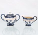 Açucareiro e leiteira em faiança japonesa - Imperial Ironstone Ware, N.K.T., Japão, meados do século XX. Conjunto composto por açucareiro com tampa, duas asas em voluta e pé anelar, acompanhado de leiteira com bico vertedor e alça lateral. Decoração derivada do Willow Pattern, em azul sob esmalte, com paisagem orientalista composta por salgueiro, construções, ponte, curso dágua, embarcação e aves em voo. Cercaduras geométricas em azul e sépia, com filetação dourada nos contornos. Superfície com craquelê de esmalte. Ambas as peças apresentam a marca "Imperial Ironstone Ware - N.K.T. - Made in Japan", associada à produção da Nihon Koshitsu Toki, atual NIKKO, manufatura japonesa fundada em Kanazawa em 1908. Medidas açucareiro: Alt.: 12,5 x Comp.: 16 x Larg.: 11cm; Medidas cremeira: Alt.: 8 x Comp.: 13 x Larg.: 9 cm