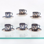 Conjunto de xícaras para café em faiança japonesa, Imperial Ironstone Ware, N.K.T., Japão, meados do século XX. Decoração derivada do Willow Pattern, em azul sob esmalte, com paisagem orientalista constituída por arquitetura, ponte, arvoredo, curso d'água, embarcação e aves em voo; cercaduras geométricas em azul e sépia e filetação dourada. Superfície com craquelê de esmalte. No reverso, marca "Imperial Ironstone Ware - N.K.T. - Made in Japan", vinculada à produção da Nihon Koshitsu Toki, atual NIKKO, manufatura fundada em Kanazawa em 1908. . Medidas xícara: Alt.: 4,5 x Comp.: 6,5 x Larg.: 4,5 cm; Medida pires: Diam.: 9,5 cm.