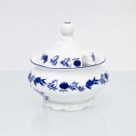 Porcelana Schmidt, Santa Catarina, Brasil (Fundada em 1945). Açucareiro em porcelana nacional, esmaltagem vítrea na cor branca, corpo bojudo moldado com gomos suaves em relevo, borda revirada para fora, tampa campaniforme encimada por puxador com feitio de fruto, dotada de abertura lateral para passagem de colher. A ornamentação consiste em decalcomania monocromática sob o vidrado em tom azul-cobalto, reproduzindo o padrão clássico saxão Zwiebelmuster (padrão cebola), composta por uma faixa contínua de romãs, pêssegos, flores estilizadas e peônias ramificadas dispostas no bojo e na borda superior da tampa, conhecida pelo senso comum como padrão "cebolinha". Marcas da manufatura ao fundo. Medidas: Alt.: 12 x Diam.: 12 cm.