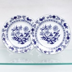 Porcelana Schmidt, Santa Catarina, Brasil (Fundada em 1945). Dois(02) pratos rasos em porcelana nacional, esmaltagem vítrea na cor branca, borda contornadas por filete azul contínuo, Orla do pires circundada por cercadura perimetral ornamentada por ramagens relevadas na própria pasta. A ornamentação consiste em decalcomania monocromática sob o vidrado em tom azul-cobalto, reproduzindo o padrão clássico saxão Zwiebelmuster (padrão cebola), composta por uma faixa contínua de romãs, pêssegos, flores estilizadas e peônias ramificadas dispostas internamente e externamente, e na orla do pires. Padrão conhecido pelo senso comum como "cebolinha". Marcas da manufatura no verso. Medida: Diam.: 26 cm.