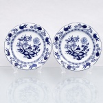 Porcelana Schmidt, Santa Catarina, Brasil (Fundada em 1945). Dois(02) pratos para sobrmesa em porcelana nacional, esmaltagem vítrea na cor branca, borda contornadas por filete azul contínuo, Orla do pires circundada por cercadura perimetral ornamentada por ramagens relevadas na própria pasta. A ornamentação consiste em decalcomania monocromática sob o vidrado em tom azul-cobalto, reproduzindo o padrão clássico saxão Zwiebelmuster (padrão cebola), composta por uma faixa contínua de romãs, pêssegos, flores estilizadas e peônias ramificadas dispostas internamente e externamente, e na orla do pires. Padrão conhecido pelo senso comum como "cebolinha". Marcas da manufatura no verso. Medida: Diam.: 20 cm.