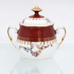 Porcelana Schmidt, Santa Catarina, Brasil (Fundada em 1945). Açucareiro em porcelana nacional, c.1950-60. Corpo bojudo de borda recortada em ondulações, base anelar e alças laterais em voluta revestida por douramento. A ornamentação exibe faixa superior em esmalte vermelho-bordeaux com cercadura de gregas em ouro, enquanto a seção inferior e a tampa de fundo branco ostentam guirlandas florais policromadas ladeadas por ornatos dourados. Marca da manufatura ao fundo. Medidas: Alt.: 12,5 x Comp.: 14,5 x Larg.: 10,5 cm.