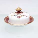 Porcelana Schmidt, Santa Catarina, Brasil (Fundada em 1945). Manteigueira coberta em porcelana nacional, c.1950-60. Conjunto composto por prato base circular com rebaixo central e tampa em formato de cúpula semiesférica com pomo cônico dourado. A aba do prato exibe esmalte vermelho-bordeaux com cercadura de gregas em ouro, tampa de fundo branco ostenta guirlandas policromadas de rosas, violetas e miosótis ladeadas por ornatos dourados. Marca da manufatura no verso. Medidas: Alt.: x Diam.: