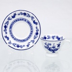 Porcelana Schmidt, Santa Catarina, Brasil (Fundada em 1945). Chávena para café em porcelana nacional, esmaltagem vítrea na cor branca, bojo hemisférico assentado sobre base circular elevada em pedestal, asa lateral modelada em forma de voluta, bordas contornadas por filete azul contínuo. Orla do pires circundada por cercadura perimetral ornamentada por ramagens relevadas na própria pasta. A ornamentação consiste em decalcomania monocromática sob o vidrado em tom azul-cobalto, reproduzindo o padrão clássico saxão Zwiebelmuster (padrão cebola), composta por uma faixa contínua de romãs, pêssegos, flores estilizadas e peônias ramificadas dispostas internamente e externamente, e na orla do pires. Padrão conhecido pelo senso comum como "cebolinha". Marcas da manufatura ao fundo.  Medidas: Xícara: Alt.: 5 x Comp.: 9 x Larg.: 7 cm; Pires: Diam.: 11cm.