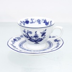 Porcelana Schmidt, Santa Catarina, Brasil (Fundada em 1945). Chávena para café em porcelana nacional, esmaltagem vítrea na cor branca, bojo hemisférico assentado sobre base circular elevada em pedestal, asa lateral modelada em forma de voluta, bordas contornadas por filete azul contínuo. Orla do pires circundada por cercadura perimetral ornamentada por ramagens relevadas na própria pasta. A ornamentação consiste em decalcomania monocromática sob o vidrado em tom azul-cobalto, reproduzindo o padrão clássico saxão Zwiebelmuster (padrão cebola), composta por uma faixa contínua de romãs, pêssegos, flores estilizadas e peônias ramificadas dispostas internamente e externamente, e na orla do pires. Padrão conhecido pelo senso comum como "cebolinha". Marcas da manufatura ao fundo.  Medidas: Xícara: Alt.: 5 x Comp.: 9 x Larg.: 7 cm; Pires: Diam.: 11cm.