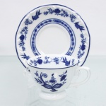 Porcelana Schmidt, Santa Catarina, Brasil (Fundada em 1945). Chávena para café em porcelana nacional, esmaltagem vítrea na cor branca, bojo hemisférico assentado sobre base circular elevada em pedestal, asa lateral modelada em forma de voluta, bordas contornadas por filete azul contínuo. Orla do pires circundada por cercadura perimetral ornamentada por ramagens relevadas na própria pasta. A ornamentação consiste em decalcomania monocromática sob o vidrado em tom azul-cobalto, reproduzindo o padrão clássico saxão Zwiebelmuster (padrão cebola), composta por uma faixa contínua de romãs, pêssegos, flores estilizadas e peônias ramificadas dispostas internamente e externamente, e na orla do pires. Padrão conhecido pelo senso comum como "cebolinha". Marcas da manufatura ao fundo.  Medidas: Xícara: Alt.: 5 x Comp.: 9 x Larg.: 7 cm; Pires: Diam.: 11cm.