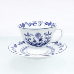 Porcelana Schmidt, Santa Catarina, Brasil (Fundada em 1945). Chávena para chá em porcelana nacional, esmaltagem vítrea na cor branca, bojo hemisférico assentado sobre base circular elevada em pedestal, asa lateral modelada em forma de voluta, bordas contornadas por filete azul contínuo. Orla do pires circundada por cercadura perimetral ornamentada por ramagens relevadas na própria pasta. A ornamentação consiste em decalcomania monocromática sob o vidrado em tom azul-cobalto, reproduzindo o padrão clássico saxão Zwiebelmuster (padrão cebola), composta por uma faixa contínua de romãs, pêssegos, flores estilizadas e peônias ramificadas dispostas internamente e externamente, e na orla do pires. Padrão conhecido pelo senso comum como "cebolinha". Marcas da manufatura ao fundo. Medidas: Xícara: Alt.: 7 x Comp.: 11,5 x Larg.: 9,5cm; Pires: Diam.: 14,5 cm.