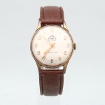 Relógio de pulso Mondaine De Luxe, fabricação suíça, circa 1960-1970. Relógio mecânico de corda manual, com movimento de 17 rubis, conforme inscrição "Ancre 17 Rubis" no mostrador. Caixa circular de metal dourado, mostrador em tonalidade champanhe com algarismos arábicos aplicados, escala perimetral de minutos e três ponteiros. Assinado "Mondaine De Luxe" e "T Swiss Made T". Fundo em aço inoxidável, marcado "Shockprotected", "Antimagnetic", "Stainless Steel Back" e "Swiss Made". Pulseira em material sintético marrom. Apresenta sinais de uso e desgaste no acabamento. Não está funcionando. Medida da caixa: Diam.:  31mm.