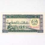Numismática / Cédulas Estrangeiras. Cédula de 100 Kip, emitida pela República Democrática Popular do Laos, referência Pick P-30a. Anverso com composição alusiva à produção agrícola, representando trabalhadores durante o processamento de cereais com máquina debulhadora, tendo ao fundo arquitetura religiosa, palmeiras e torres de transmissão de energia; emblema estatal no campo superior. Reverso com caminhões atravessando ponte sobre curso d'água, povoado e soldado armado junto a instalações de armazenamento. Marca-d'água composta por foice e martelo com estrelas. Impressão policromática sobre papel fiduciário. Laos, s.d., emissão de 1979-1988.