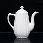 Porcelana Real, Mauá - SP(Fundada em 1943 por Fritz Erwin Schmidt). Bule para café em porcelana nacional, esmaltagem vítrea na cor branca, corpo cilíndrico, aproximadamente cônico, moldado com sucessão de caneluras em relevo, bico vertedor sinuoso de ponta recortada, tampa ligeiramente abualada encimada por pomo de contorno losangular, alça latral estilizada relevada com folhagens acantiformes. Marca da manufatura ao fundo. Medidas: Alt.: 23,5 x Comp.: 23,5 x Larg.: 12 cm.