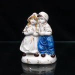 Grupo figurativo em porcelana, cxomposição moldada representando casal sentado em banco, disposto em atitude de aproximação, com as cabeças inclinadas entre si. Figura feminina portando vestido e touca, mãos reunidas à frente; personagem masculino trajando camisa, barrete e calças em azul. Policromia sob e sobre o vidrado em azul, castanho, ocre e vermelho, com realces a ouro nas vestes, cabelos, calçados e elementos da base. Plinto integrado, de contorno irregular, com pequenos motivos vegetalistas dourados. Medidas: Alt.: 11,5 x Comp.: 8,5 x Larg.: 5,5cm.