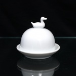 Pequena cloche para porção individual de manteiga ou patê de fígado de pato(foie gras) executado em porcelana branca vitrificada, datável de meados do século XX. O conjunto apresenta estrutura dividida em duas seções independentes com acabamento liso e esmalte brilhante de tom uniforme. A base é constituída por pires circular provido de cavidade interna rebaixada para o encaixe da guarnição, ladeada por aba externa curva de perfil suave. A cobertura superior apresenta formato hemiesférico ou cúpula lisa, encimada por puxador moldado em formato tridimensional de ave aquática repousada. Medidas: Alt.: 9 x Diam.: 9,5 cm.