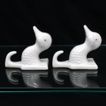 Par de ganchos de parede em porcelana, esmaltação branca, modelados em forma de patos, com cabeça e pescoço projetados em relevo, plumagem indicada por escamas moldadas e olhos pontuados em preto. Cada exemplar integra placa posterior de contorno retangular, provida de dois orifícios para fixação em parede, prolongando-se inferiormente em gancho recurvado. Medidas: Alt.: 9 x Comp.: 6 x Larg.: 9 cm.