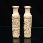Par de vasos(bud vase) executados em marfim oriental entalhado, datável do período da Dinastia Qing, circa final do século XIX. Apresentam corpo tubular, colo estrangulado e boca circular de bordo evertido, delimitados por frisos torneados. Painel central exibe a representação mítica de dragões imperiais de cinco garras que serpenteiam por entre nuvens estilizadas (xiangyun) e folhagens densas, capturando a expressividade das criaturas com olhos destacados por pigmentação escura, composição recorrente na iconografia oriental e documentada em peças de marfim do século XIX. Apresentam perda da base de madeira original. Medidas: Alt.: 13 x Diam.: 3 cm.