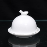 Pequena cloche para porção individual de manteiga ou patê de fígado de pato(foie gras) executado em porcelana branca vitrificada, datável de meados do século XX. O conjunto apresenta estrutura dividida em duas seções independentes com acabamento liso e esmalte brilhante de tom uniforme. A base é constituída por pires circular provido de cavidade interna rebaixada para o encaixe da guarnição, ladeada por aba externa curva de perfil suave. A cobertura superior apresenta formato hemiesférico ou cúpula lisa, encimada por puxador moldado em formato tridimensional de ave. Medidas: Medidas: Alt.: 7,5 x Diam.: 9,5 cm.