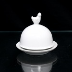 Pequena cloche para porção individual de manteiga ou patê de fígado de pato(foie gras) executado em porcelana branca vitrificada, datável de meados do século XX. O conjunto apresenta estrutura dividida em duas seções independentes com acabamento liso e esmalte brilhante de tom uniforme. A base é constituída por pires circular provido de cavidade interna rebaixada para o encaixe da guarnição, ladeada por aba externa curva de perfil suave. A cobertura superior apresenta formato hemiesférico ou cúpula lisa, encimada por puxador moldado em formato tridimensional de ave. Medidas: Medidas: Alt.: 7,5 x Diam.: 9,5 cm.Medidas xícara: Alt.: 9 x Diam.: 9,5cm.