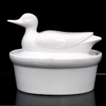 Terrina em cerâmica esmaltada branca, com tampa figurativa em forma de pato(duck tureen). Recipiente de formato oval, paredes lisas e bordo moldurado; cobertura ajustada, modelada integralmente como ave aquática em repouso, com bico alongado, cabeça erguida, asas e plumagem definidas em baixo-relevo. Superfície revestida por esmalte branco de acabamento brilhante. Na base, etiqueta comercial parcialmente preservada com inscrições "MICR... e ...WASHER SAFE", indicando fabricação destinada ao uso culinário em micro-ondas e à lavagem em máquina.  Medidas: Alt.: 19 x Comp.: 23,5 x Larg.: 17,5 cm.