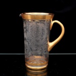 Jarra executada em cristal europeu, circa 1930-1950. Corpo cilíndrico alongado com bico vertedor e alça lateral arqueada. Superfície integralmente ornamentada por gravação fosca, apresentando composição de inspiração oriental, com reservas reticuladas, volutas, folhagens, aves em voo, elementos arquitetônicos e paisagem com vegetação. Bordo, friso inferior e alça recobertos por douradura aplicada. Base circular de fundo espesso. Medidas: Alt.: 21 x Comp.: 17 x Larg.: 12,5 cm.