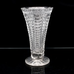 Vaso para flores executado em cristal translúcido e incolor. Apresenta corpo em formato obcônico que se expande em direção à boca, sustentado por pedestal curto sobre base circular; superfície externa decorada com sucessão de gomos horizontais moldados em relevo, organizados em colunas verticais, borda superior com acabamento lapidado em pequenos ovais sequenciais, base circular com fundo preenchido por estrela de raios concêntricos.Medidas: Alt.: 20,5 x Diam.: 11cm.