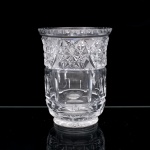 Vaso executado em cristal incolor lapidado. Corpo cilíndrico de paredes espessas, assente sobre base circular moldurada. Decoração distribuída em registros horizontais: faixa superior composta por estrelas multipontas executadas por cortes prismáticos, intercaladas por reservas triangulares preenchidas por quadriculado de "ponta de diamante"; segmento intermediário articulado por painéis verticais, facetas ogivais e incisões geométricas; porção inferior lavrada com cortes lenticulares, losangos e motivos radiais. Bordo espesso, exteriormente recortado por sucessão de festões semicirculares. Fundo com estrela radial lapidada. Provavelmente Europa, circa 19001930. Medidas: Alt.: 20,5 x Diam.: 15,5 cm.