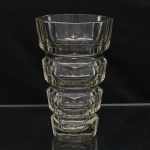 Design atribuído a Josef Hoffmann (18701956) para Moser, Karlsbad (Karlovy Vary), Tchecoslováquia. Vaso executado em cristal incolor, de inspiração Art Déco, circa 1930-1950. Corpo de formato obcônico facetado com oito lados, estruturado em quatro registros horizontais escalonados, de diâmetros progressivamente reduzidos em direção à base. Cada seção apresenta paredes espessas, definidas por planos facetados e arestas chanfradas, estabelecendo sucessão de volumes geométricos sobrepostos. Boca de contorno octogonal, com bordo biselado, acompanhando a articulação prismática do corpo. Termina em base maciça, igualmente facetada. Medidas: Alt.: x Diam.: